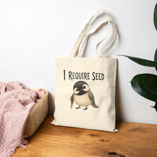 A cream canvas tote bag with long handles, featuring a black text graphic that reads 'I REQUIRE SEED' above a small chickadee illustration. The bag is styled on a wooden table next to a pink blanket in a basket and a green plant, against a white wall.
