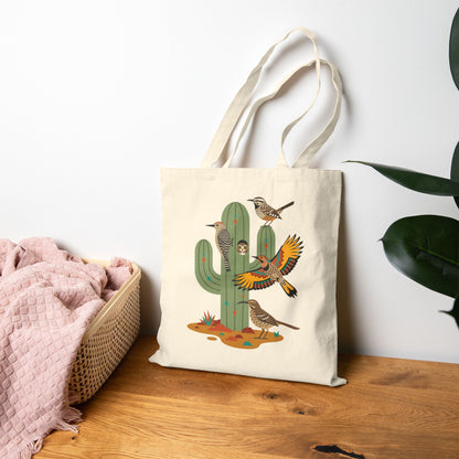 A beige canvas tote bag with a cactus and bird design sitting on a table with a blanket and plant. 