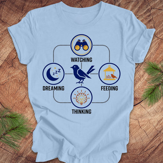 Light blue t-shirt with a bird graphic and "dreaming, watching, feeding, thinking" text and graphics on a wood background.