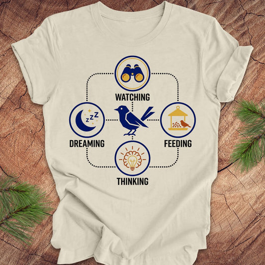 Beige t-shirt with a bird graphic and "dreaming, watching, feeding, thinking" text and graphics on a wood background.