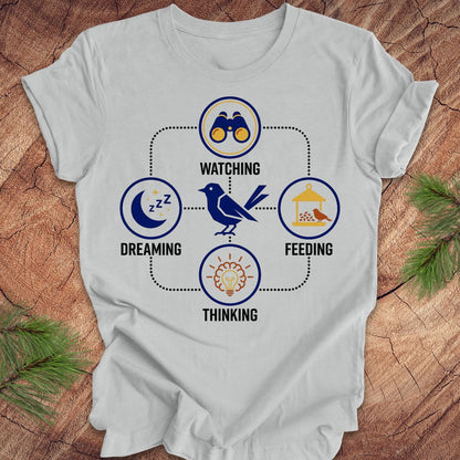 Gray t-shirt with a bird graphic and "dreaming, watching, feeding, thinking" text and graphics on a wood background.