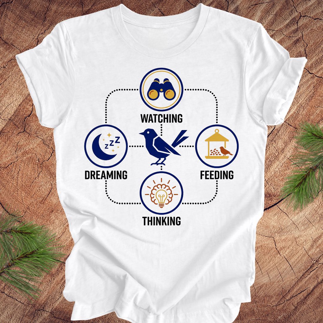 White t-shirt with a bird graphic and "dreaming, watching, feeding, thinking" text and graphics on a wood background.