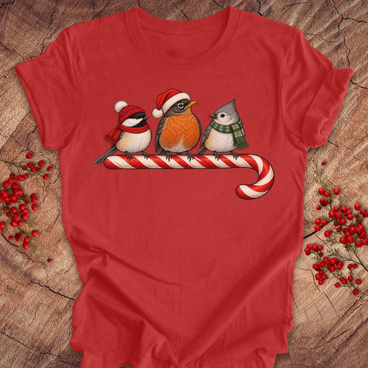 Heather red t-shirt with a Black-capped Chickadee, American Robin, and Tufted Titmouse in winter hats and scarves perched on a candy cane, laid flat on a rustic wood background.