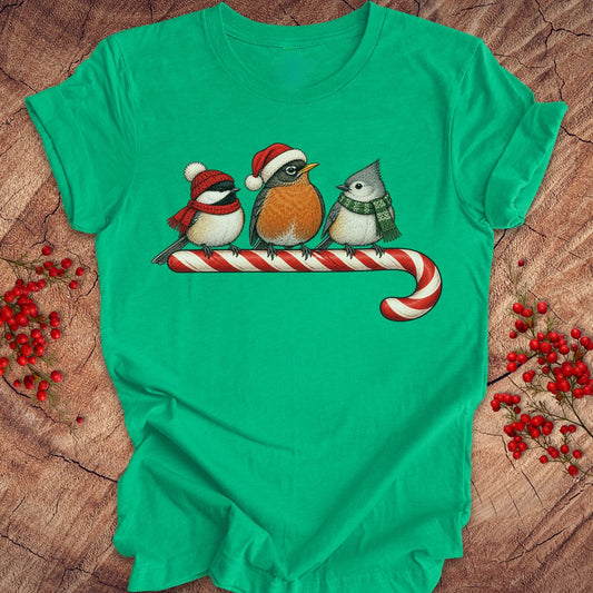 Heather kelly green t-shirt with a Black-capped Chickadee, American Robin, and Tufted Titmouse in winter hats and scarves perched on a candy cane, laid flat on a rustic wood background.