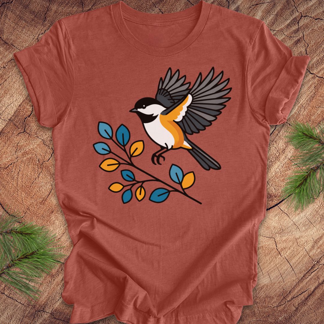 A terracotta-colored short-sleeve t-shirt featuring a graphic of a chickadee bird perched on a branch with blue, yellow, and orange leaves, laid flat on a rustic wood background with pine needles.