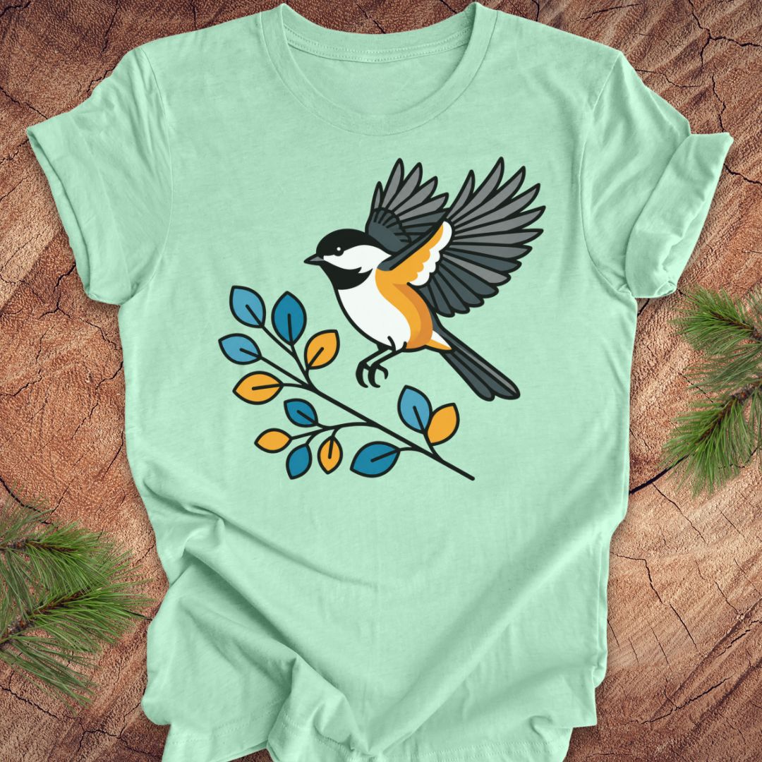 A mint green short-sleeve t-shirt laid flat on a rustic wood background, featuring a graphic of a chickadee bird perched on a branch with blue and yellow leaves.
