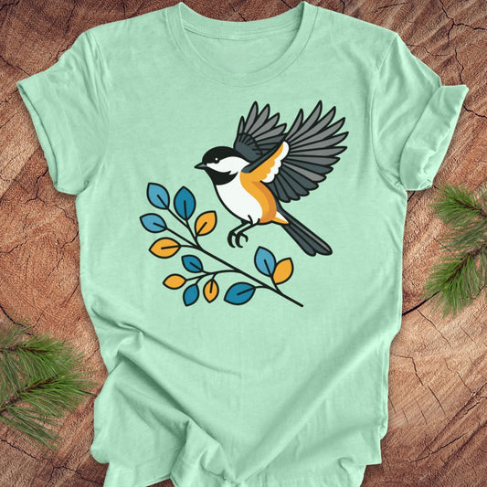 A mint green short-sleeve t-shirt laid flat on a rustic wood background, featuring a graphic of a chickadee bird perched on a branch with blue and yellow leaves.