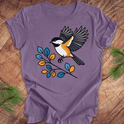 A purple short-sleeve crewneck t-shirt featuring a graphic of a chickadee bird perched on a branch with blue, yellow, and orange leaves, laid flat on a rustic wood background with pine needles.