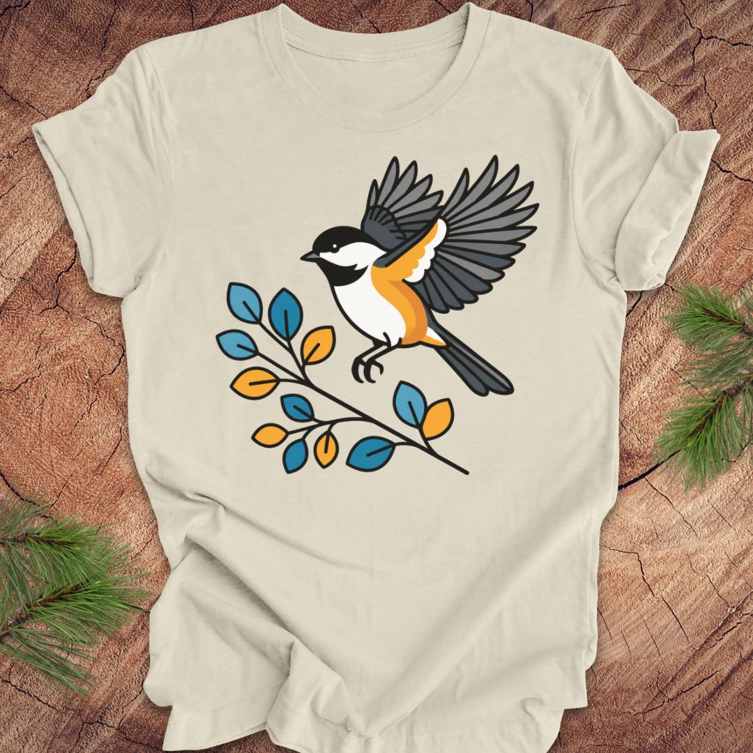 A light beige short-sleeve t-shirt featuring a colorful graphic of a chickadee bird perched on a branch with blue and yellow leaves, laid flat on a rustic wood background with pine needles in the corners.