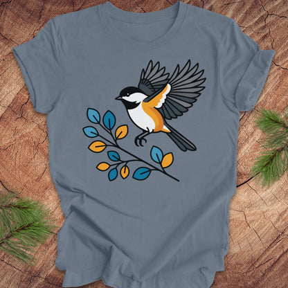 A slate blue crewneck t-shirt featuring a graphic of a chickadee bird perched on a branch with blue, yellow, and orange leaves, laid flat on a rustic wood background with pine needles in the corners.
