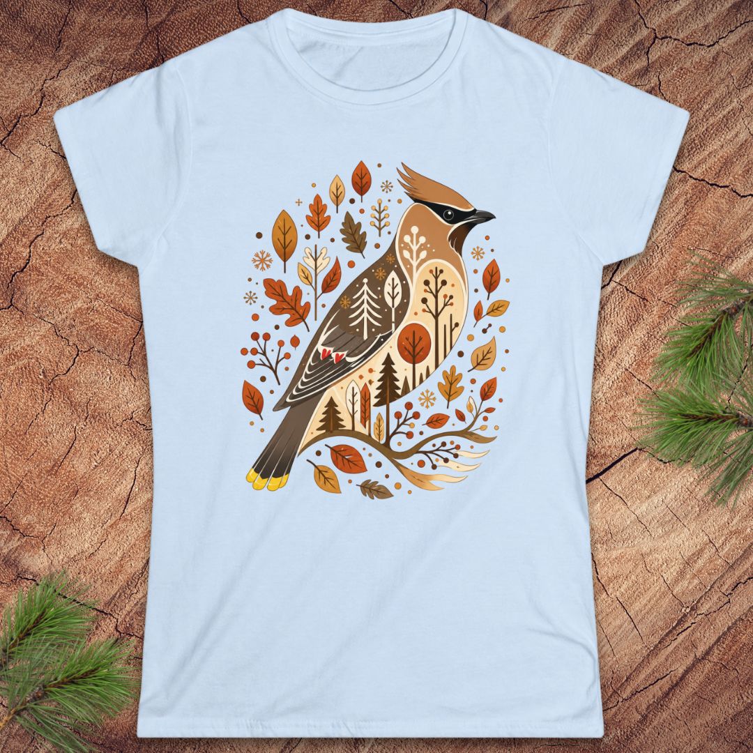 A light blue short-sleeve crewneck t-shirt featuring a detailed graphic of a cedar waxbird surrounded by autumn leaves and trees, laid flat on a rustic wood background with pine branches in the corners.