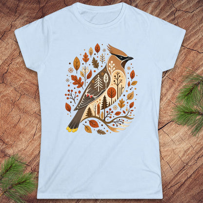 A light blue short-sleeve crewneck t-shirt featuring a detailed graphic of a cedar waxbird surrounded by autumn leaves and trees, laid flat on a rustic wood background with pine branches in the corners.