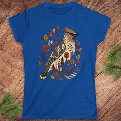 A royal blue short-sleeve t-shirt featuring a detailed graphic of a cedar waxwing bird surrounded by autumn leaves and trees, laid flat on a rustic wood background with pine branches in the corners.