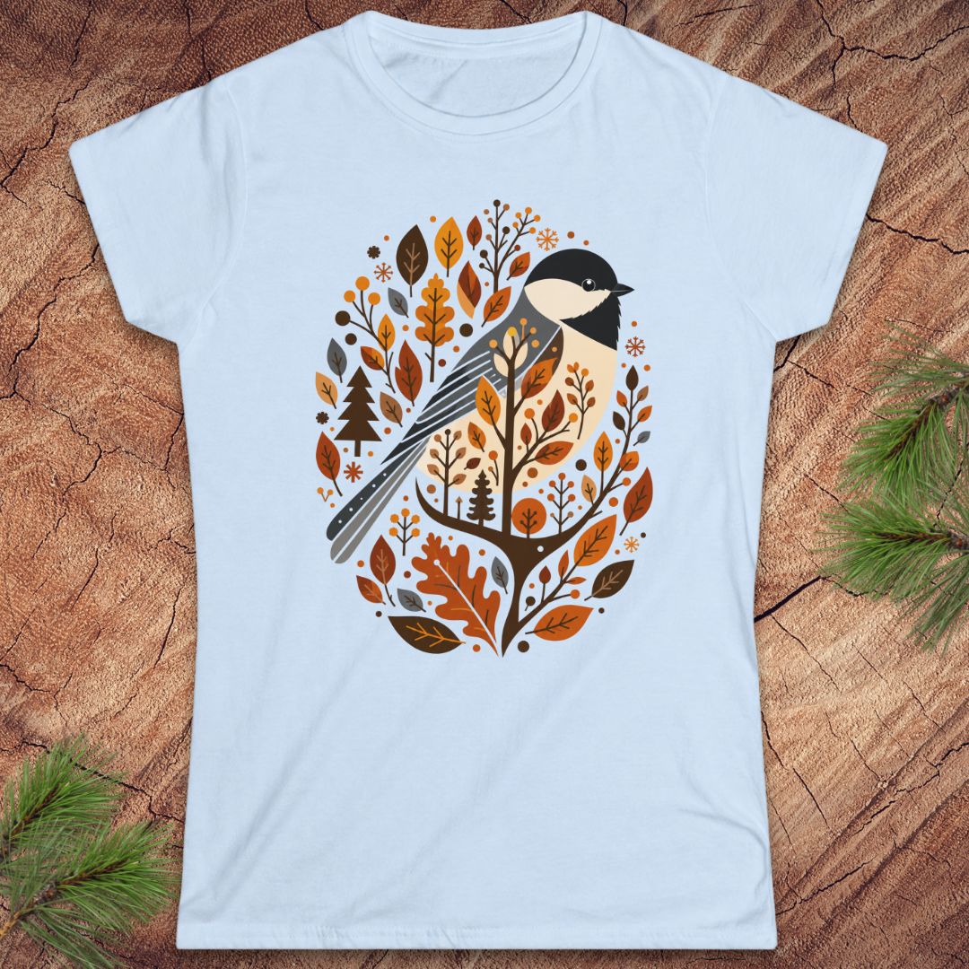 A light blue short-sleeve crewneck t-shirt featuring a stylized bird and autumn leaves graphic, laid flat on a rustic wood background with pine branches in the corners.