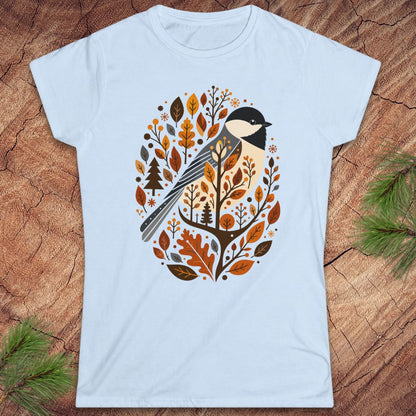 A light blue short-sleeve crewneck t-shirt featuring a stylized bird and autumn leaves graphic, laid flat on a rustic wood background with pine branches in the corners.