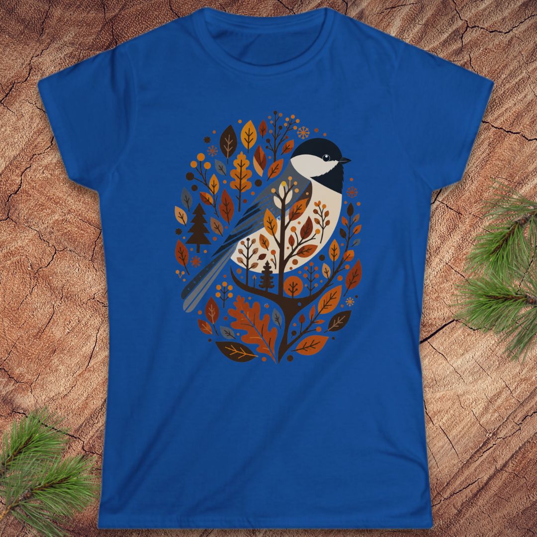 A royal blue short-sleeve crewneck t-shirt featuring a stylized graphic of a bird perched among autumn leaves, laid flat on a rustic wood background with pine branches in the corners.