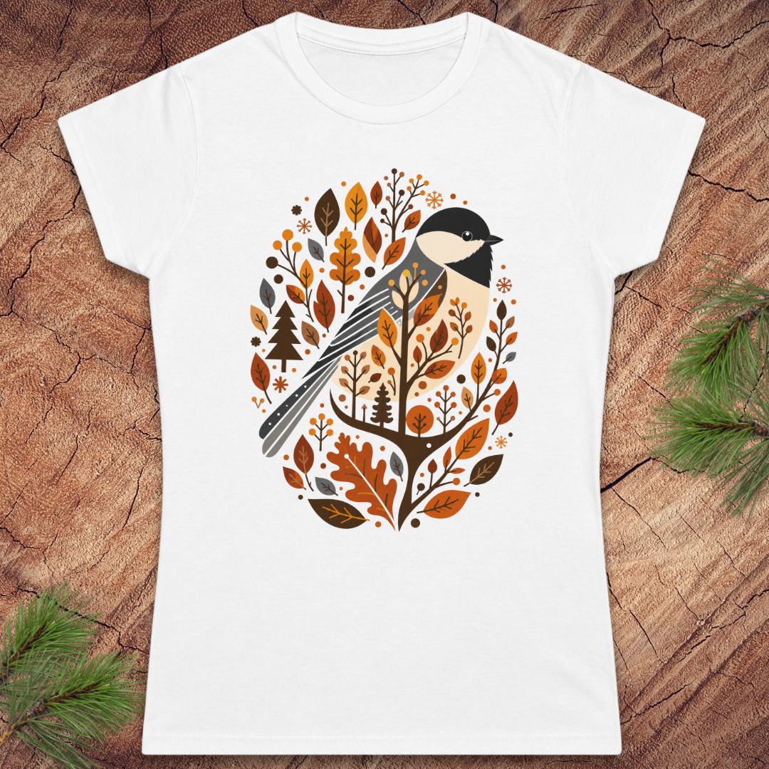 A white short-sleeve crewneck t-shirt featuring a stylized graphic of a bird surrounded by autumn leaves and trees, displayed flat on a rustic wood background with pine branches in the corners.