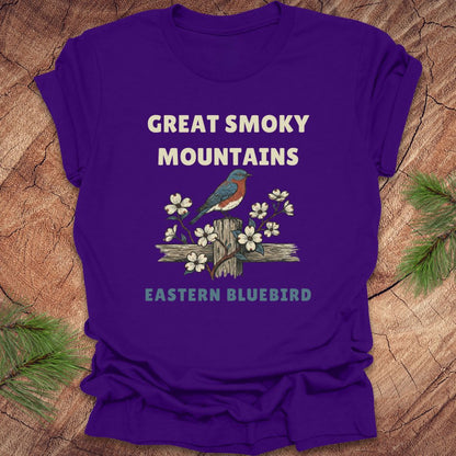 Purple t-shirt with an illustration of an Eastern Bluebird and text on a wooden background