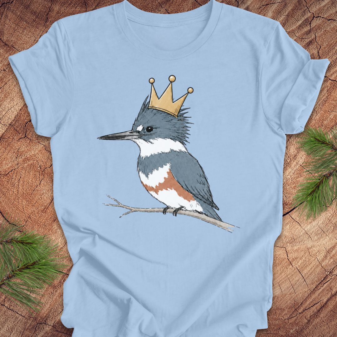 A light blue short-sleeve crewneck t-shirt featuring a graphic of a blue-and-white kingfisher wearing a gold crown, displayed flat on a rustic wood background with pine needles.