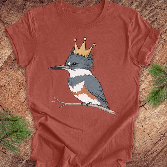 A rust-colored short-sleeve t-shirt featuring a graphic of a blue-gray and white kingfisher wearing a gold crown, displayed flat on a rustic wood background with pine needles.