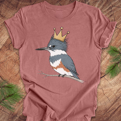A dusty rose short-sleeve t-shirt featuring a graphic of a blue-gray and white kingfisher wearing a gold crown, laid flat on a rustic wood background with pine needles in the corners.