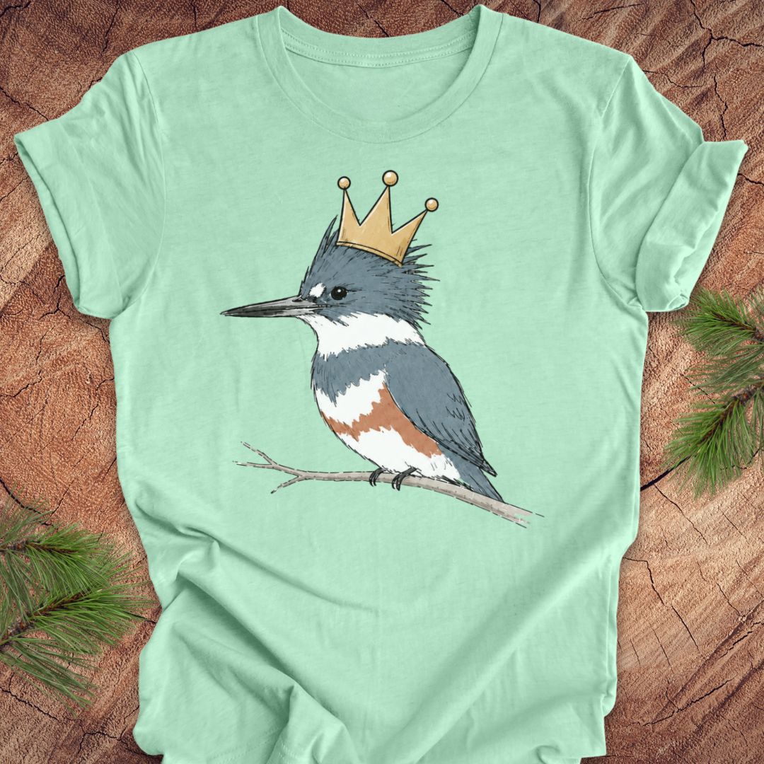 A mint green short-sleeve t-shirt laid flat on a rustic wood surface, featuring a printed illustration of a kingfisher wearing a gold crown.