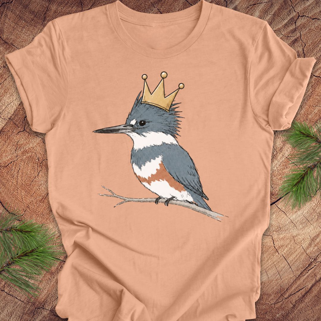 A peach-colored short-sleeve t-shirt featuring a graphic of a blue-gray and white kingfisher wearing a gold crown, displayed flat on a rustic wood background with pine branches.