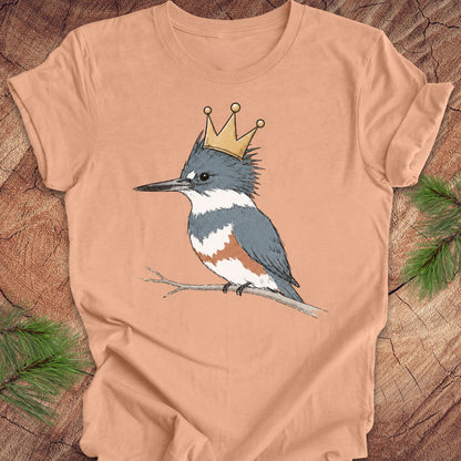 A peach-colored short-sleeve t-shirt featuring a graphic of a blue-gray and white kingfisher wearing a gold crown, displayed flat on a rustic wood background with pine branches.