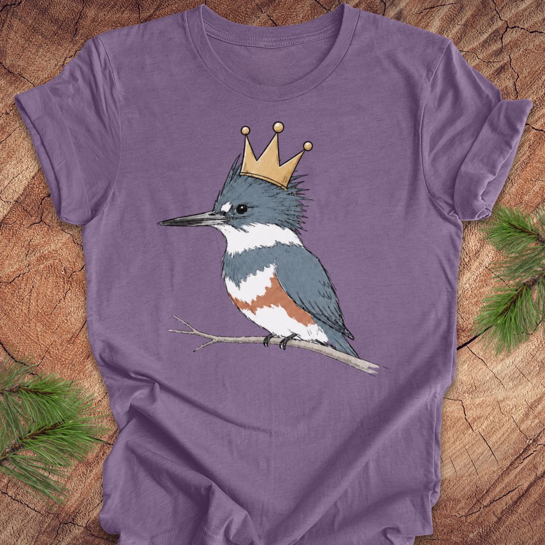 A purple short-sleeve t-shirt featuring a graphic of a blue-gray and white kingfisher wearing a gold crown, displayed flat on a rustic wood background with pine branches.