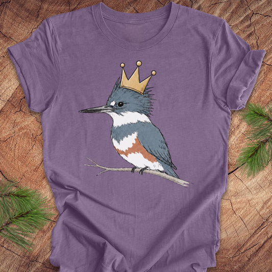 A purple short-sleeve t-shirt featuring a graphic of a blue-gray and white kingfisher wearing a gold crown, displayed flat on a rustic wood background with pine branches.