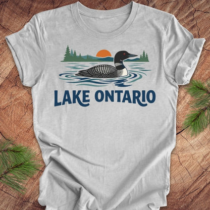 A light gray short-sleeve t-shirt with a graphic of a loon in Lake Ontario, featuring pine trees, a sunset, and the text 'LAKE ONTARIO' in blue. The shirt is displayed flat on a rustic wood background with pine branches in the corners.
