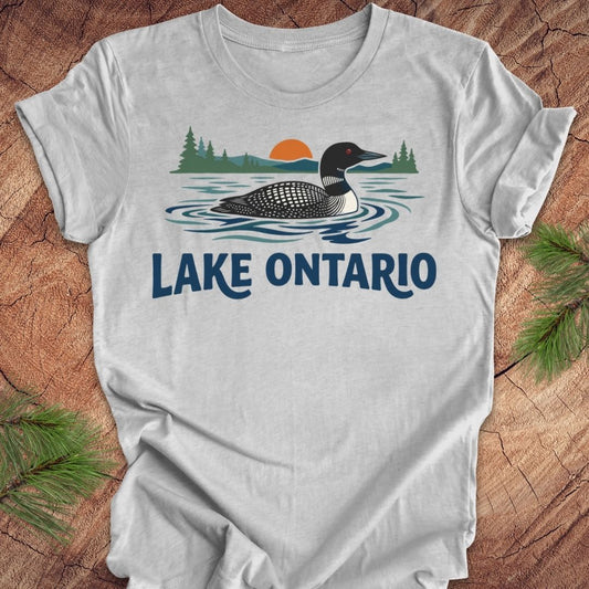 A light gray short-sleeve t-shirt with a graphic of a loon in Lake Ontario, featuring pine trees, a sunset, and the text 'LAKE ONTARIO' in blue. The shirt is displayed flat on a rustic wood background with pine branches in the corners.
