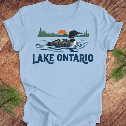 A light blue short-sleeve t-shirt laid flat on a rustic wood surface, featuring a graphic of a loon in Lake Ontario with pine trees, a sunset, and the text 'LAKE ONTARIO' in dark blue. Green pine branches are placed in the corners.