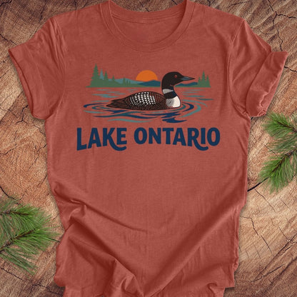 A terracotta-colored short-sleeve t-shirt featuring a graphic of a loon on Lake Ontario with a sunset and trees, laid flat on a rustic wood background with pine branches.
