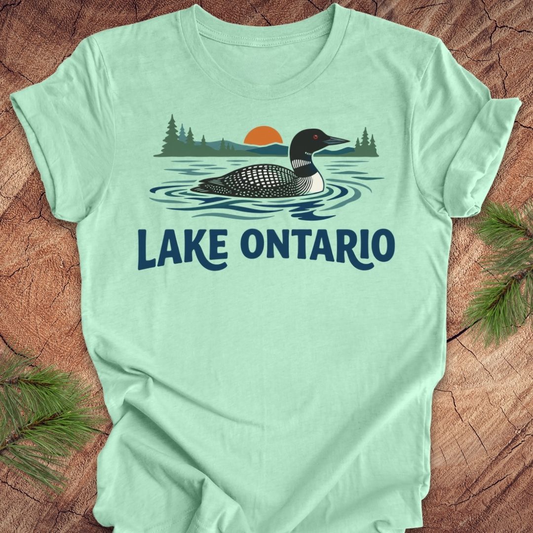 A mint green short-sleeve t-shirt laid flat on a rustic wood background, featuring a graphic of a loon in Lake Ontario with pine trees and an orange sunset, and the text 'LAKE ONTARIO' printed below.