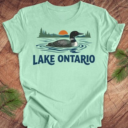 A mint green short-sleeve t-shirt laid flat on a rustic wood background, featuring a graphic of a loon in Lake Ontario with pine trees and an orange sunset, and the text 'LAKE ONTARIO' printed below.