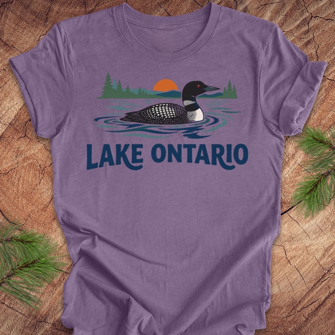 A purple short-sleeve t-shirt laid flat on a rustic wood background with pine branches in the corners. The shirt features a graphic of a loon in Lake Ontario with a sunset and trees, and the text 'LAKE ONTARIO' printed below.