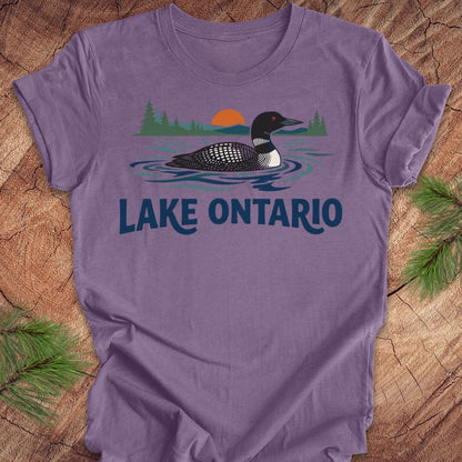 A purple short-sleeve t-shirt laid flat on a rustic wood background with pine branches in the corners. The shirt features a graphic of a loon in Lake Ontario with a sunset and trees, and the text 'LAKE ONTARIO' printed below.