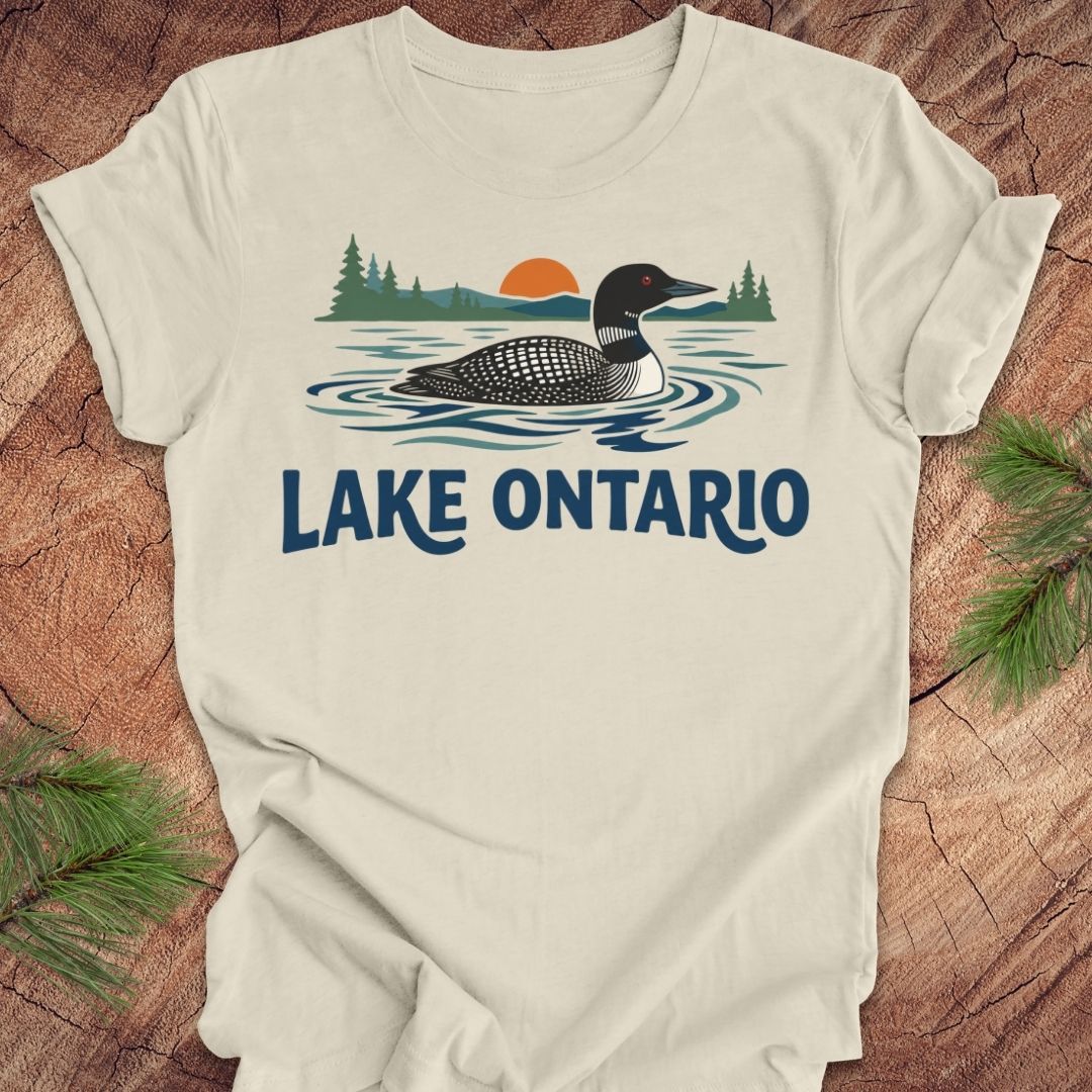 An off-white t-shirt laid flat on a rustic wood background, featuring a graphic of a loon in Lake Ontario with pine trees and a sunset, with 'LAKE ONTARIO' printed below in blue.
