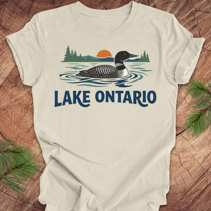 An off-white t-shirt laid flat on a rustic wood background, featuring a graphic of a loon in Lake Ontario with pine trees and a sunset, with 'LAKE ONTARIO' printed below in blue.