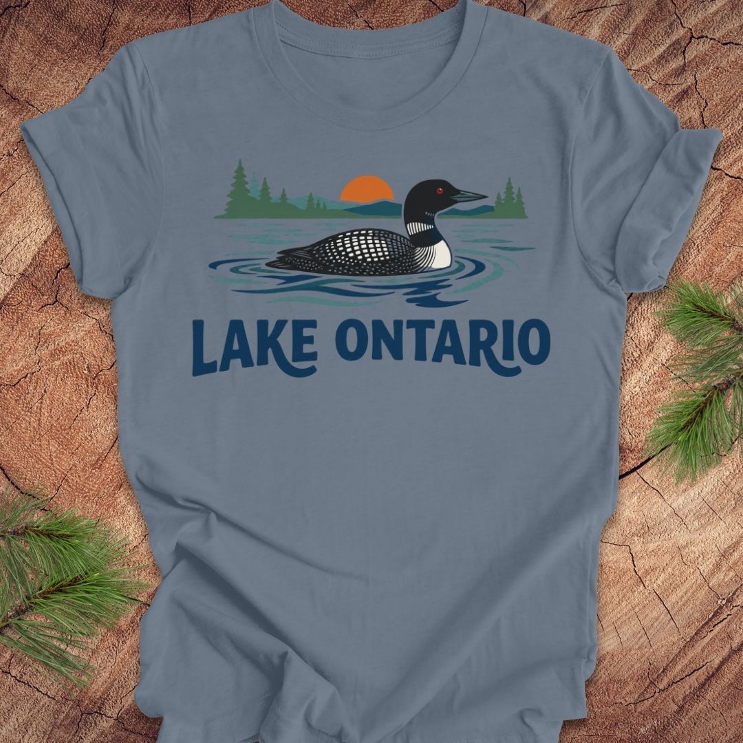 A slate blue crewneck t-shirt featuring a graphic of a loon in Lake Ontario with pine trees and an orange sunset, laid flat on a rustic wood background with pine needles.