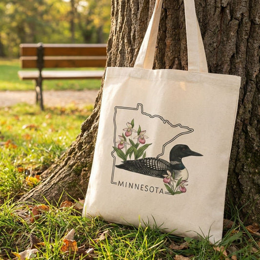 Tote bag with Minnesota loon design leaning against a tree in a park.
