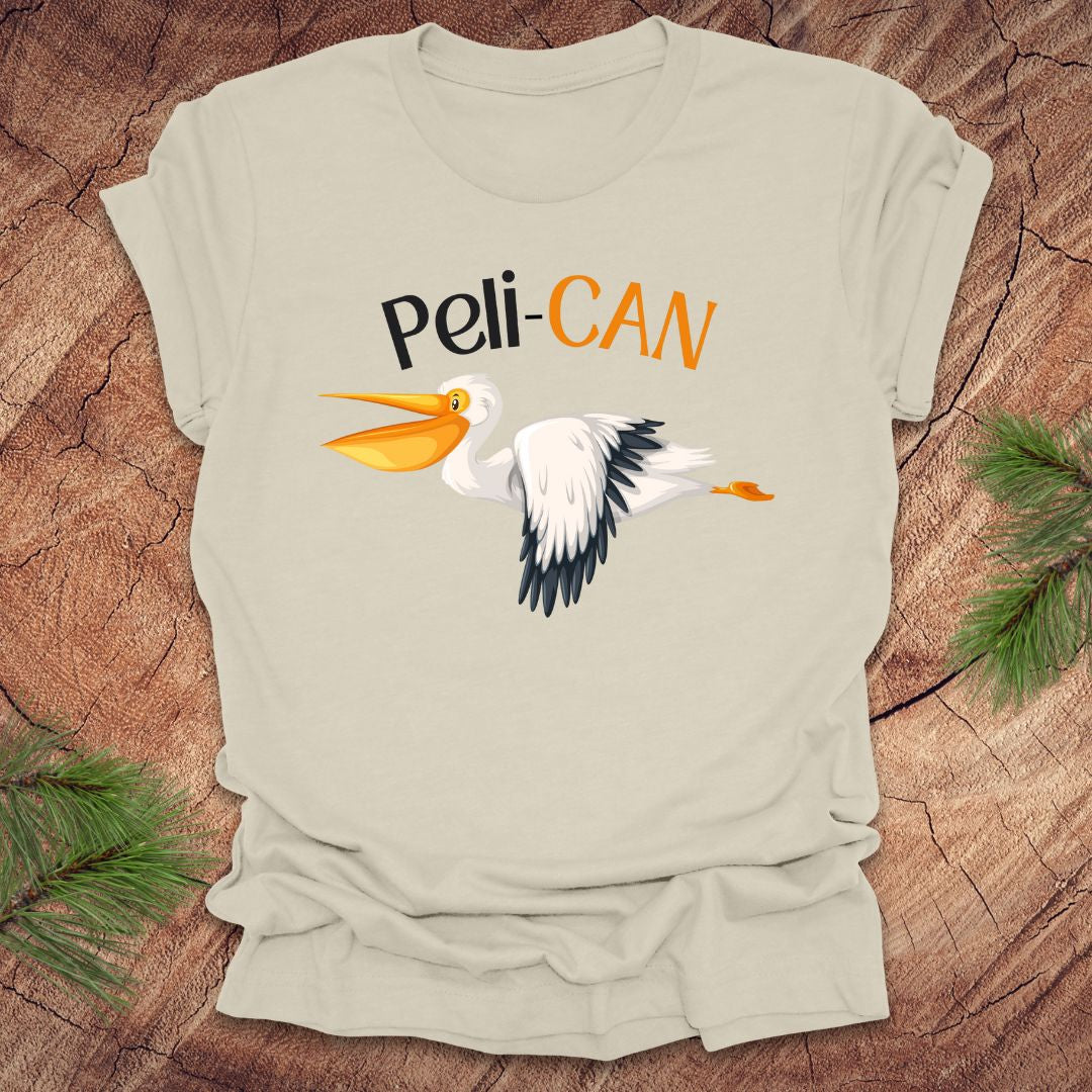 Beige t-shirt with a pelican graphic and 'Peli-CAN' text on a wooden surface.