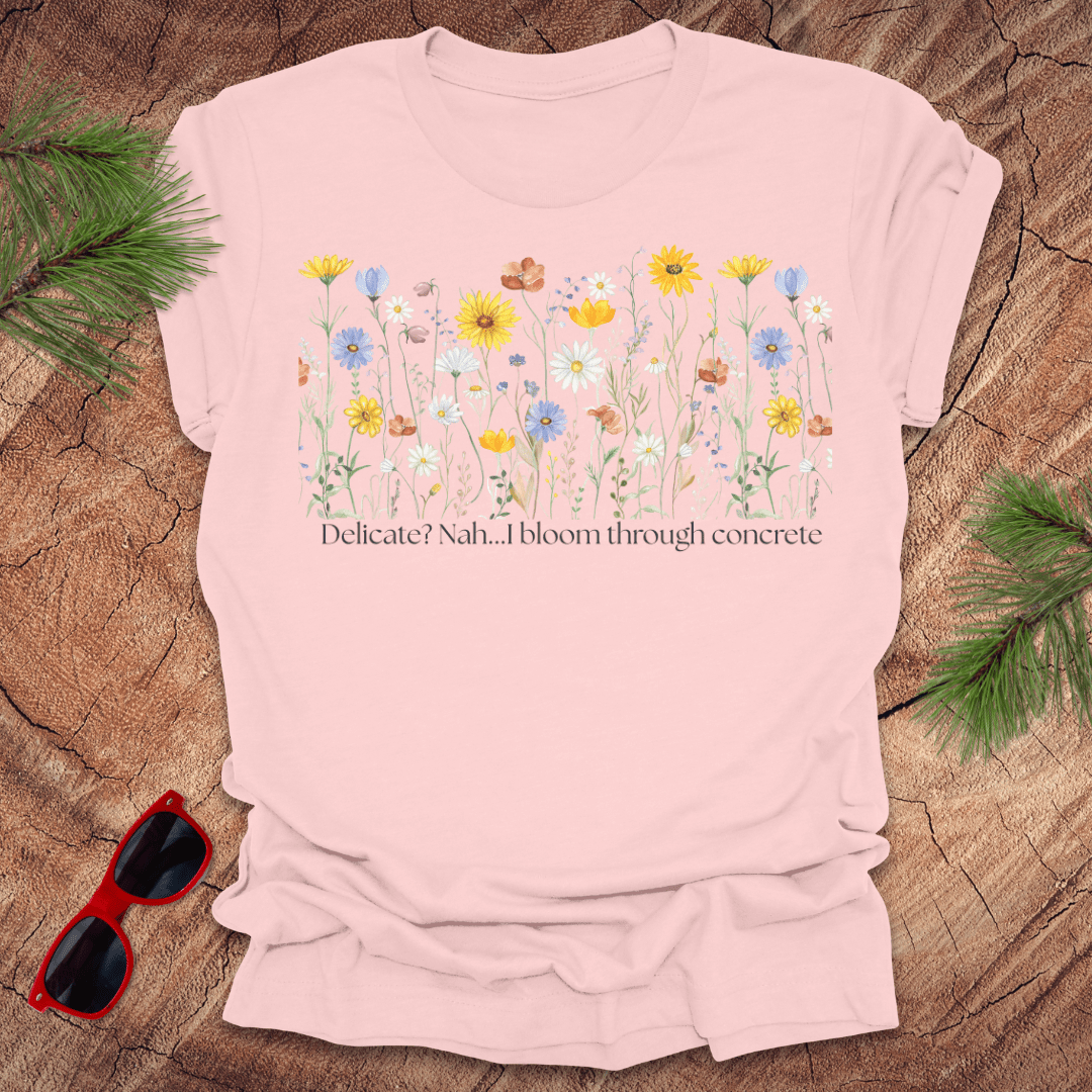 Pink t-shirt with floral design and text "delicate? nah. i bloom through concrete.' on a wooden surface.