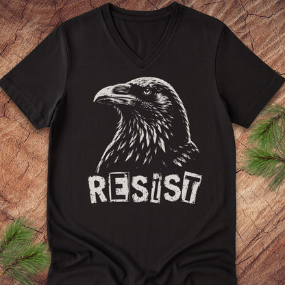 A black V-neck t-shirt featuring a white graphic of a raven's head and the word 'RESIST' in a distressed stencil font, laid flat on a rustic wood background with pine branches in the corners.