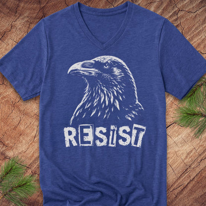 A heathered blue V-neck t-shirt featuring a distressed white graphic of a raven and the word 'RESIST', laid flat on a rustic wood background with pine needles in the corners.