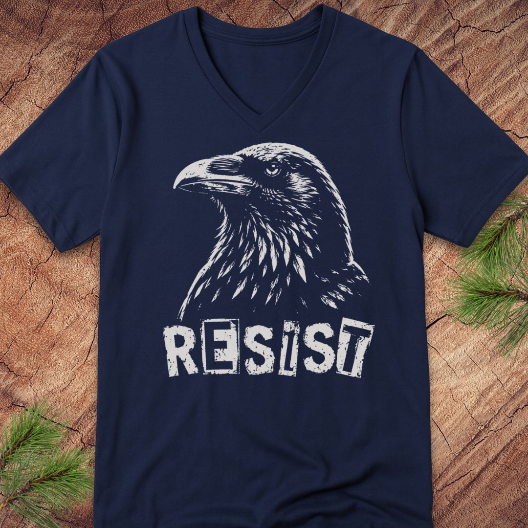 A navy blue V-neck t-shirt featuring a distressed white graphic of a raven and the word 'RESIST', laid flat on a rustic wood background with pine needles in the corners.