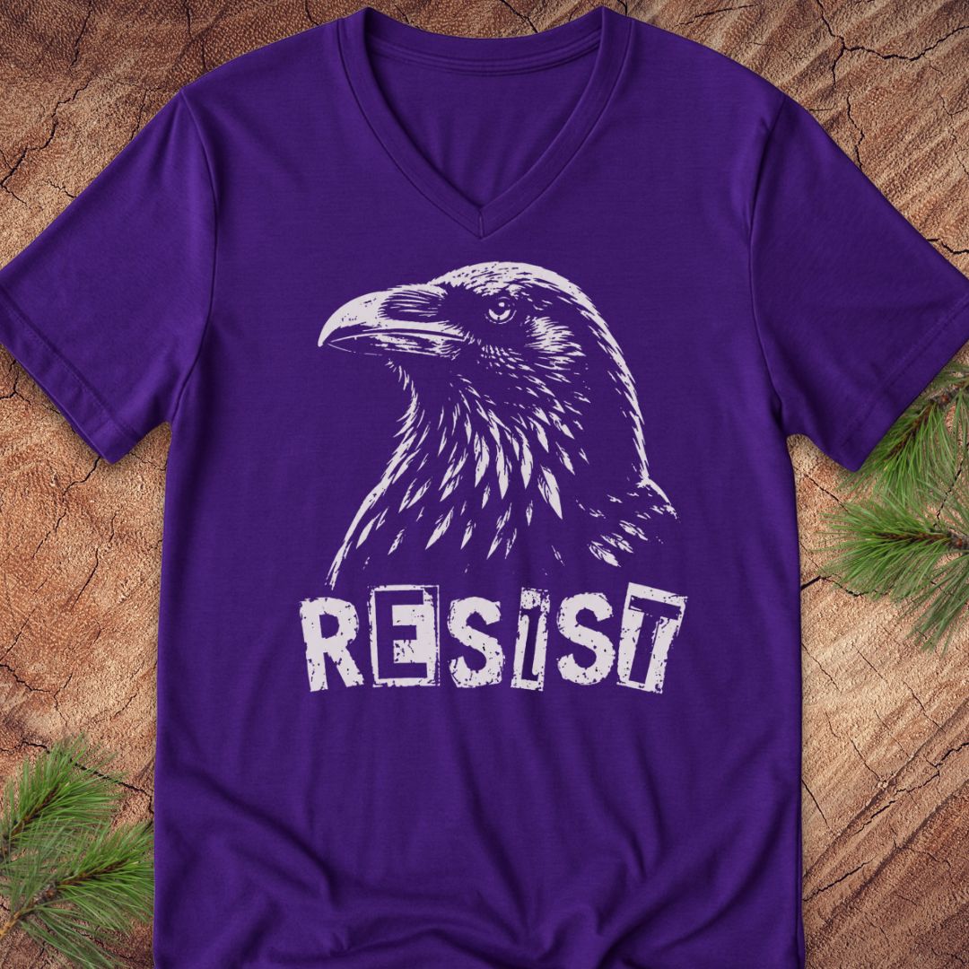 A purple V-neck t-shirt featuring a white distressed graphic of a raven and the word 'RESIST', laid flat on a rustic wood background with pine needles in the corners.