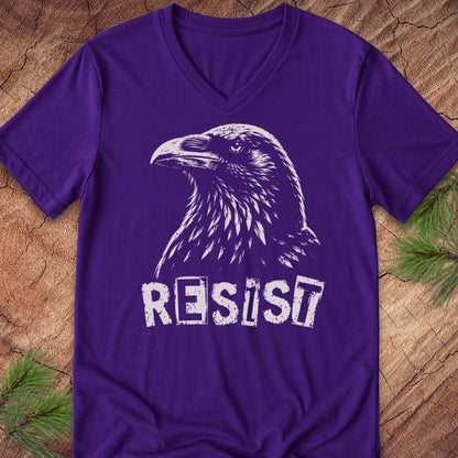 A purple V-neck t-shirt featuring a white distressed graphic of a raven and the word 'RESIST', laid flat on a rustic wood background with pine needles in the corners.