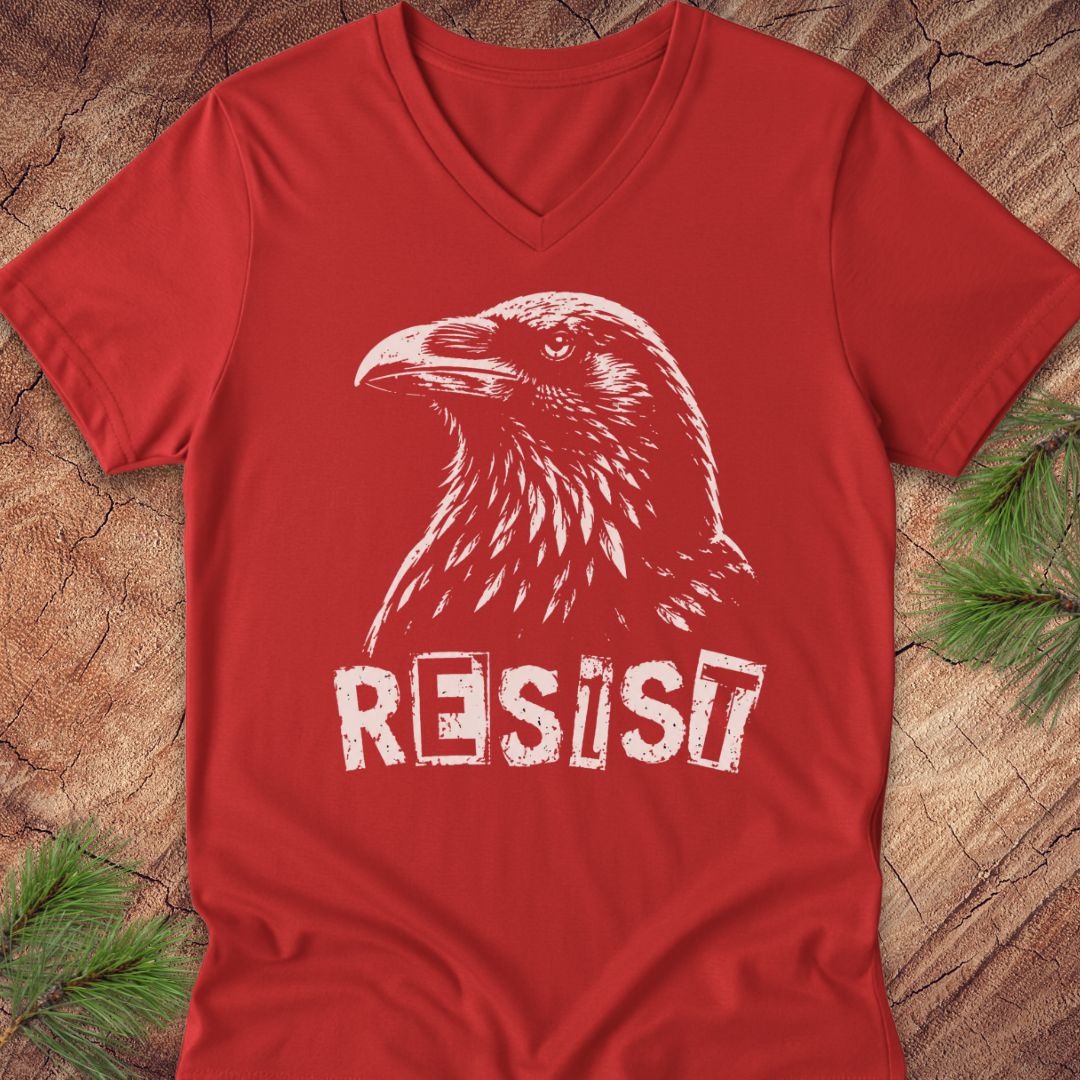 A red V-neck t-shirt featuring a white graphic of a raven and the word 'RESIST' in a distressed stencil font, laid flat on a rustic wood background with pine needles.